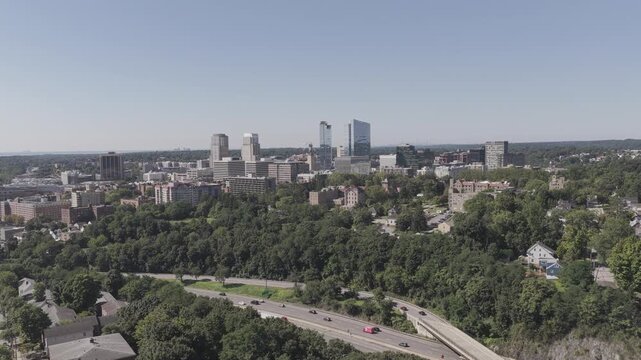 A steady drone hover frames the White Plains skyline in New York above the wooded hillside and a divided highway, with moving traffic, low-rise neighborhoods and large buildings under a clear sky.