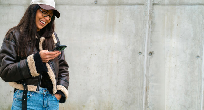 Beautiful young laughing woman with brown leather jacket and eyeglasses using her phone outdor