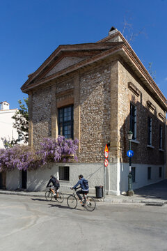 Two cyclists riding bikes on a street corner, passing a traditional stone building covered with blooming purple wisteria flowers under a clear blue sky in Athens, Greece