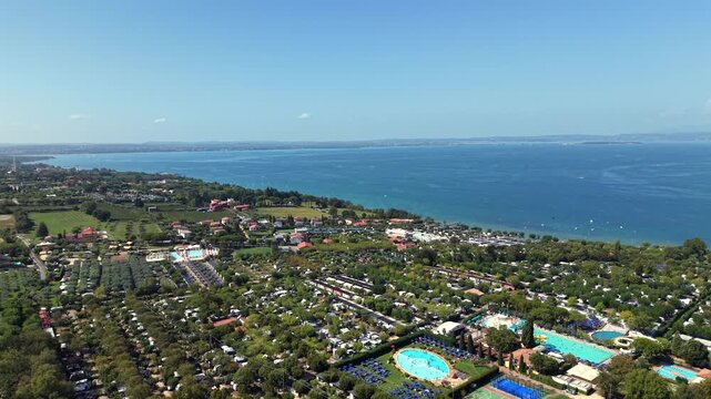 Resort campsite with turquoise pools spreads through olive groves near Lazise, Lake Garda, lakeside village and terracotta rooftops line hillside beneath clear blue sky. Aerial drone wide shot