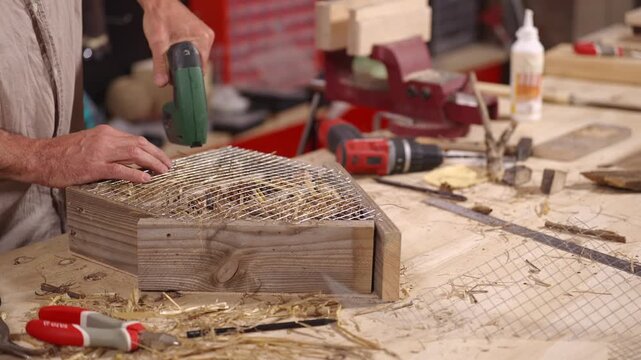 Hands secure wire mesh to a wooden insect hotel filled with natural materials using a staple gun at a workshop bench, creating shelter for garden wildlife.