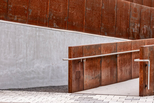 Minimalist architecture with concrete and weathered metal ramp, handrails and geometric textures shaping an exterior walkway with strong linear depth