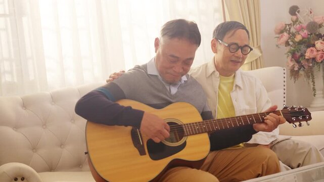 Happy Asian senior gay couple playing guitar and singing together at home. Elderly people enjoying music, leisure activity, mental wellbeing and healthy retirement lifestyle in living room.