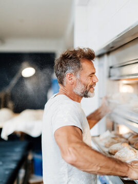 Smiling Mature Baker Taking Fresh Bread From Steaming Oven in Artisan Bakery