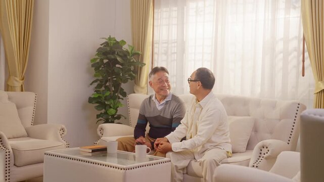 Happy Asian senior gay couple watching television together at home while relaxing with living room. Elderly lgbtq family enjoy retirement lifestyle with leisure activities and mental wellbeing