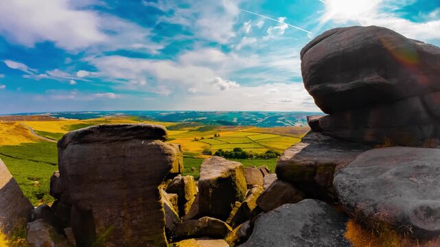 Massive weathered rock formations above green farmland and rolling hills under moving summer clouds timelapse. Rugged gritstone outcrop beside open countryside time lapse. Rural valley with road and