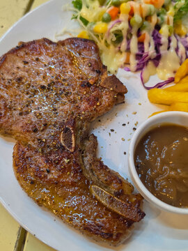 A grilled bone-in pork chop steak served with French fries, toast, brown gravy, and a creamy mixed salad of cabbage, peas, corn and lettuce.