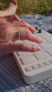 A close-up view of hands rhythmically tapping on a white electronic MIDI drum pad controller outdoors. A young woman creates electronic beats while relaxing on a picnic blanket.