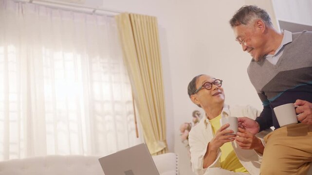 Happy Asian senior gay couple drinking coffee while reviewing income, expenses and bank statement together on laptop at home for household budgeting, financial planning and retirement money management