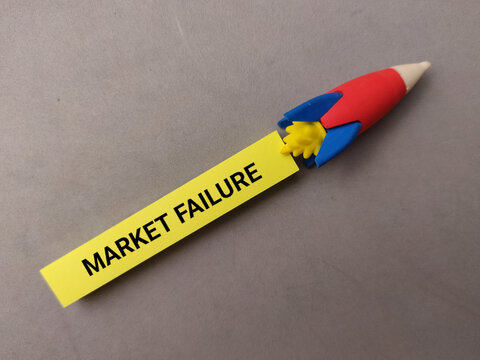 A yellow paper strip reading MARKET FAILURE is attached to a colorful toy rocket eraser on grey