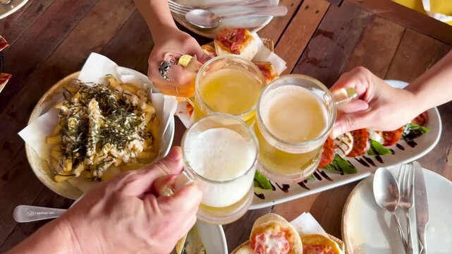 Friends clinking beer glasses together over a table full of delicious food and appetizers during a toast.