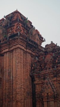 Upward camera tilt along red brick temple towers revealing carved cornices, rounded arches, pilasters, and conical spires with sculptural finials under overcast sky