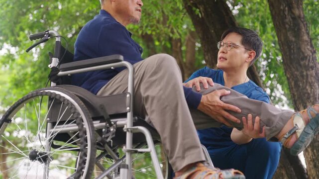 Asian male caregiver helping senior man in wheelchair stretch his leg during exercise near home. Elderly nursing health care, mobility support and physical wellbeing. Retirement home healthcare.