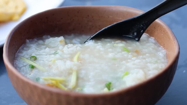 Slow motion close up of black spoon scooping hot rice porridge with ginger and spring onions in brown bowl.