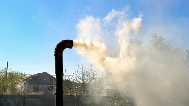 Dense gray smoke pouring from a curved metal chimney pipe on a burning barrel in a rural yard, dry grass and branches smoldering under blue sky during garden waste disposal.