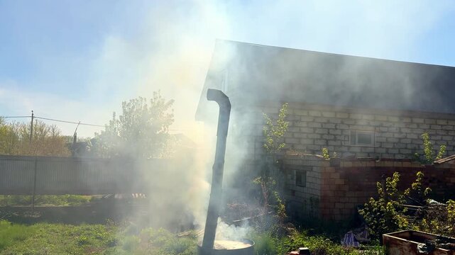 Thick white smoke rising from a bent metal pipe mounted on a burning barrel used to burn dry grass and branches in a countryside garden during autumn cleanup near an old brick house.