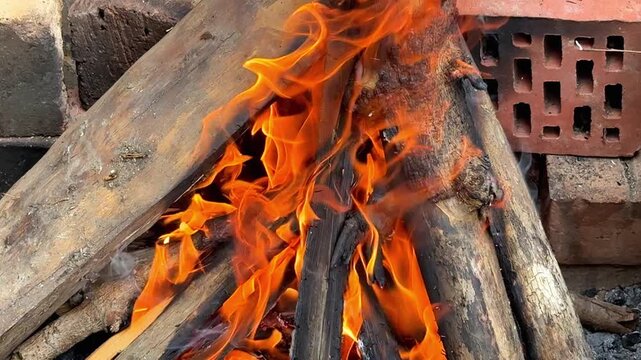 Bright orange flames burning through stacked firewood logs in an outdoor fire pit lined with bricks and stone, close up shot of a bonfire being prepared for cooking or warmth.