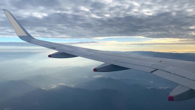 Airplane wing in flight beneath layered clouds, with sunlight breaking through gaps and illuminating the hazy landscape below.