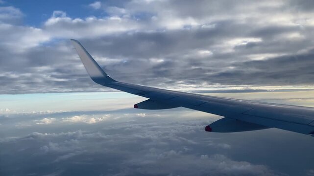 Airplane wing above scattered clouds during flight, with layered gray clouds overhead and patches of blue sky.
