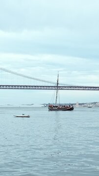 View of 25 de Abril Bridge famous tourist landmark of Lisbon connecting Lisboa and Almada wtih yacht boats passing under in cloudy weather. Lisbon, Portugal