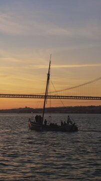 View of 25 de Abril Bridge famous tourist landmark of Lisbon connecting Lisboa and Almada on Setubal Peninsula over Tagus river with passing tourist yacht boat silhouette on sunset. Lisbon, Portugal