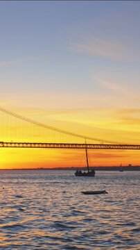 View of 25 de Abril Bridge famous tourist landmark of Lisbon connecting Lisboa and Almada on Setubal Peninsula over Tagus river with passing tourist yacht boat silhouette on sunset. Lisbon, Portugal