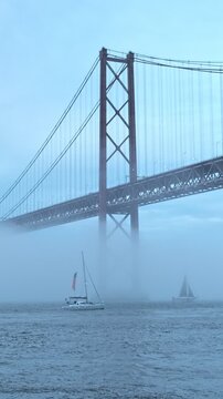 View of 25 de Abril Bridge famous tourist landmark of Lisbon connecting Lisboa and Almada in heavy fog mist with yacht boats passing under. Lisbon, Portugal