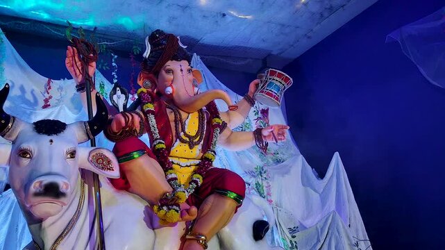 Ganesh idol dressed as Lord Shiva rides Nandi bull at night, holding trishul and damru under colorful pandal lights during Ganesh Chaturthi festival in India.
