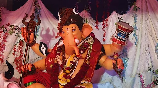 Ganesh idol dressed as Lord Shiva holds trishul with shadow silhouette on backdrop, marigold garlands decorate pandal during Ganesh Chaturthi festival India.
