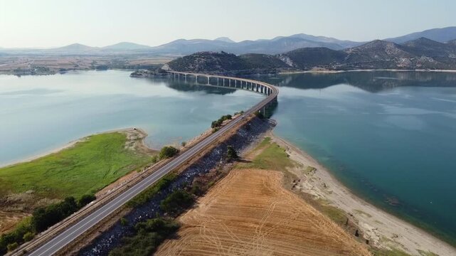 Breathtaking cinematic drone shot capturing the iconic long concrete bridge and winding road crossing the beautiful blue water reservoir of Polyfytos Lake in Kozani.