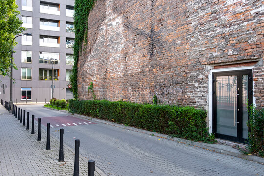 Quiet urban lane beside a weathered brick wall, hedge and residential buildings, combining pedestrian access with local city infrastructure