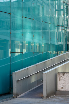 Modern access ramp beside a turquoise glass facade, combining pedestrian circulation, architecture and infrastructure within a contemporary urban setting