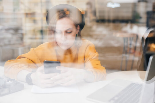 Woman using smart phone while working on laptop at cafe