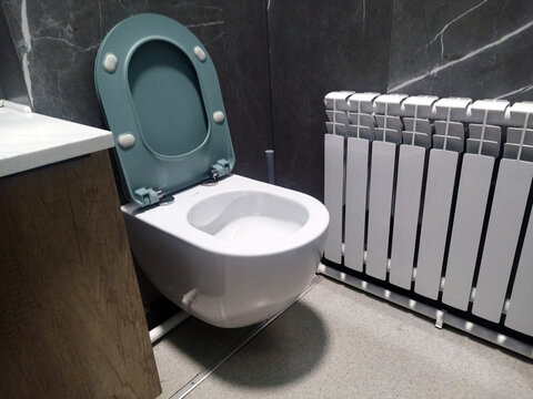 Modern white wall-hung toilet bowl with an open teal lid installed in a stylish contemporary bathroom featuring dark marble wall tiles, a wooden vanity cabinet, and a white heating radiator.