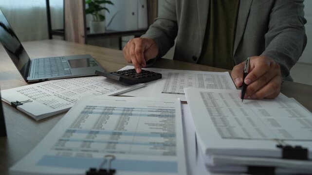 Businessman using calculator and pen with printed financial reports, reviewing expenses and revenue for annual budget planning and corporate finance analysis.
