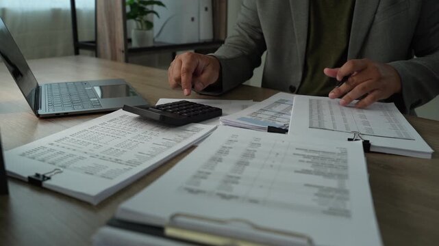 Businessman using calculator and pen with printed financial reports, reviewing expenses and revenue for annual budget planning and corporate finance analysis.