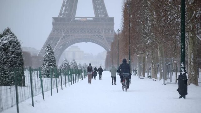 A person rides a bicycle through deep snow on the Champ de Mars in Paris, with the Eiffel Tower visible in the background. Other pedestrians walk along the snowy path during a cold winter day