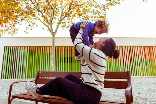 Happy mother sitting on a park bench, lifting her baby boy in the air, both smiling and having fun outdoors