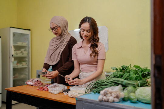 Two women work side by side preparing fresh vegetables on table in bright indoor kitchen, using knives beside leafy greens, mushrooms, cabbage, beans and assorted produce for healthy meal preparation