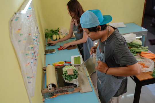 Two market workers package leafy greens and root vegetables on indoor preparation table, using plastic wrap and trays while sorting produce for retail sale in small shop workspace.