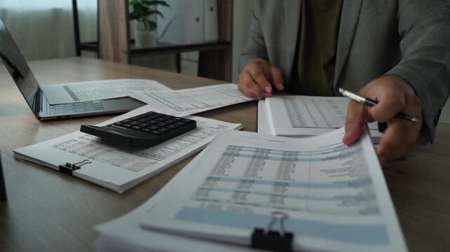 Businessman handling printed financial reports beside calculator and laptop, checking figures and documents for annual budget planning and financial review.
