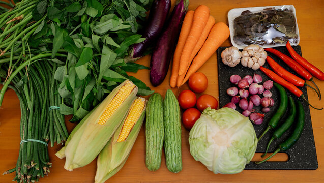 Assorted fresh vegetables on wooden table with leafy greens, carrots, eggplants, tomatoes, chilies, garlic, onions, corn, cabbage, bitter melon, beans, and herbs for healthy home cooking