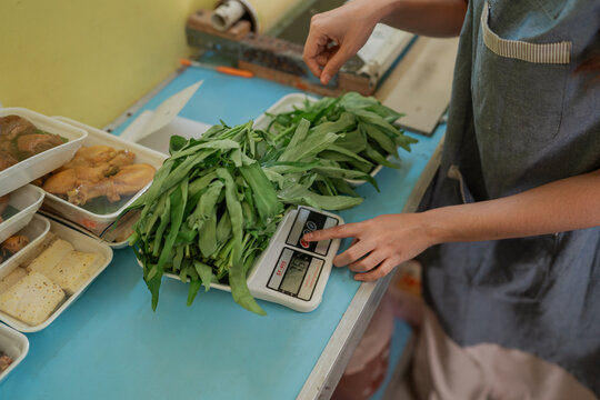 Close view of food vendor weighing fresh leafy greens on digital scale beside trays of cooked dishes and tofu at vibrant market stall, showing everyday produce sales and meal preparation.