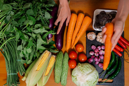 Top view of hands arranging colorful fresh vegetables on warm wooden table, including carrots, leafy greens, eggplant, corn, tomatoes, peppers, garlic, onions, cabbage, and chili peppers for meal prep