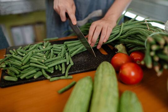 Close up of hands cutting green beans on kitchen board with tomatoes and cucumbers nearby, showing healthy fresh produce meal prep for everyday cooking.