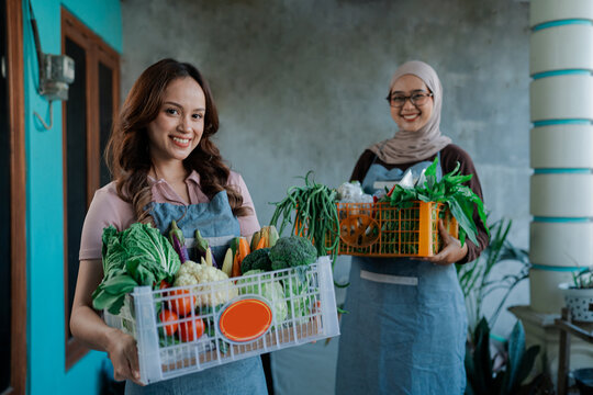 Two smiling young women stand on home porch holding crates packed with fresh vegetables. Denim aprons, one hijab, suggest local produce delivery, friendly neighborhood market teamwork.