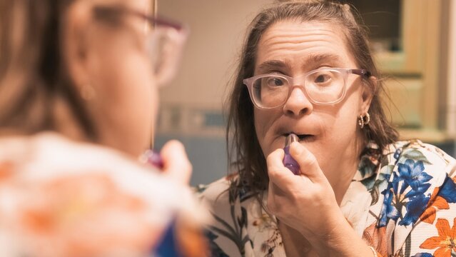 Woman with down syndrome carefully applying lipstick while looking in bathroom mirror, showcasing confidence and self-care routine