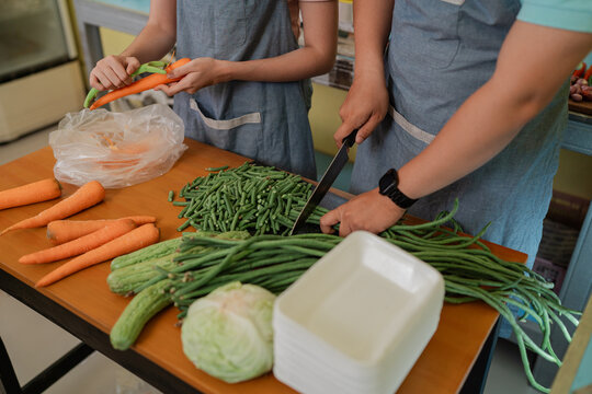 Two people in aprons sort and chop fresh carrots, green beans and cucumbers on wooden table, preparing vegetables for healthy home meal in bright indoor light.