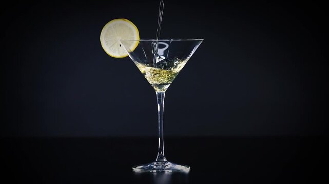 Clear martini glass with lemon slice receiving a steady stream of golden liquid forming small bubbles in a slow motion shot.