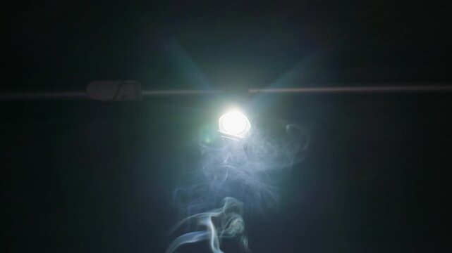 An intense light beam shines from a flashlight through floating smoke against a dark background. Fine particle diffusion creates soft volumetric rays and subtle optical flare effects.
